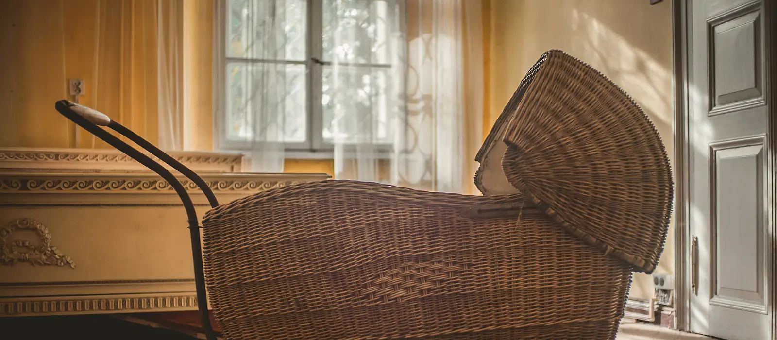 Das Bild zeigt einen Antiken Kinderwagen aus Bast, der vor einem Fenster steht.